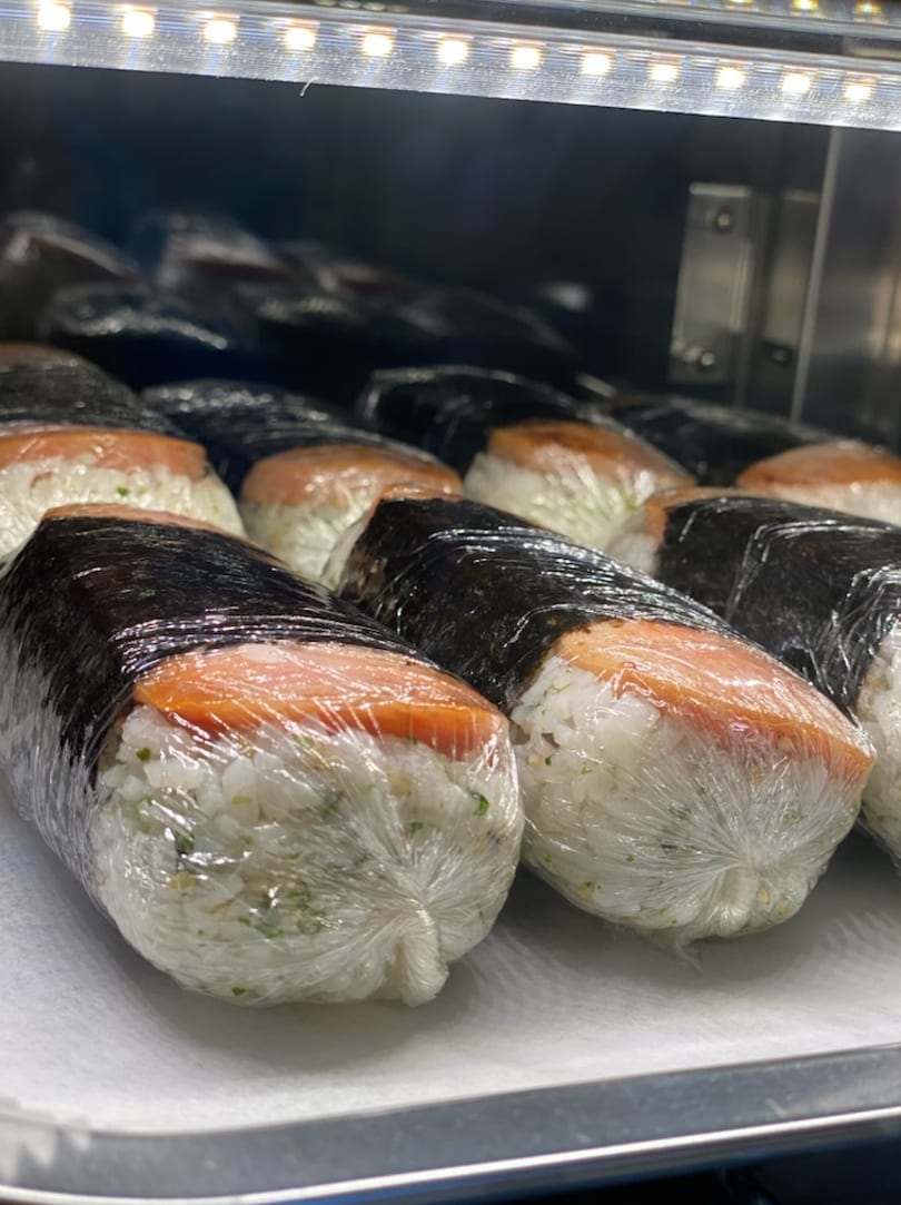 Rows of Mama Dukes Hawaiian Barbeque Spam Musubi, wrapped in plastic, under a warm light.